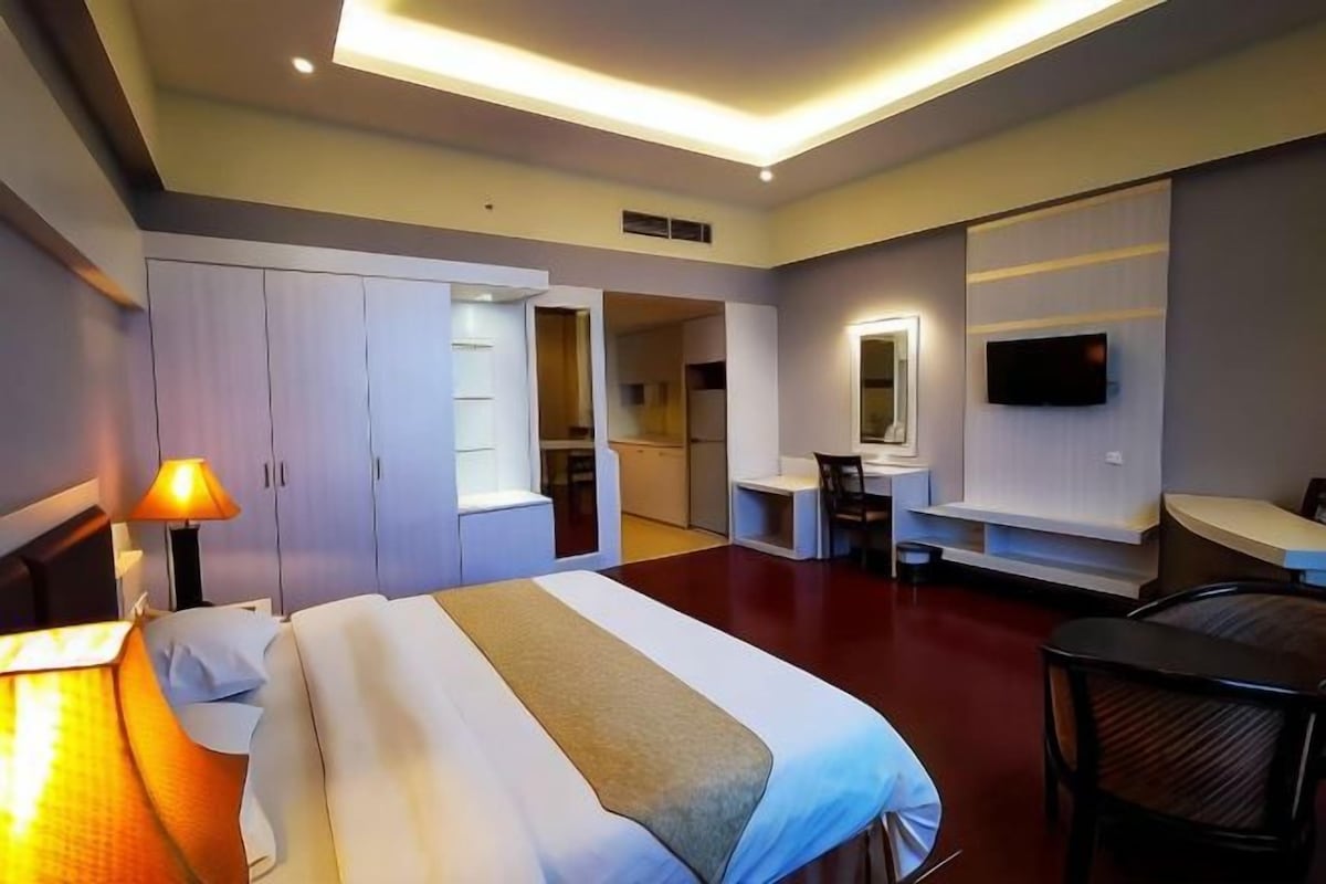A hotel room with a bed, bedside table, lamp, wardrobe, desk, chair, and a TV mounted on the wall.
