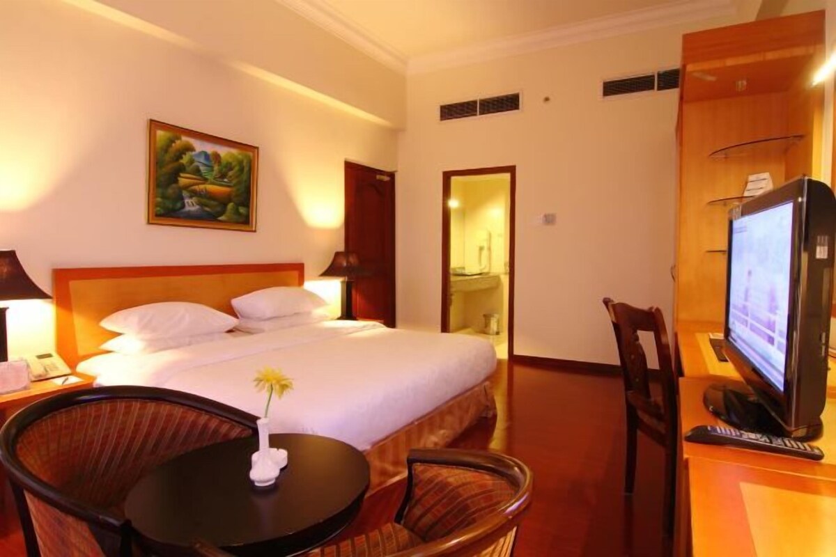 Una habitación de hotel con cama, escritorio con computadora, silla, pequeña mesa con una flor y un cuadro en la pared.
