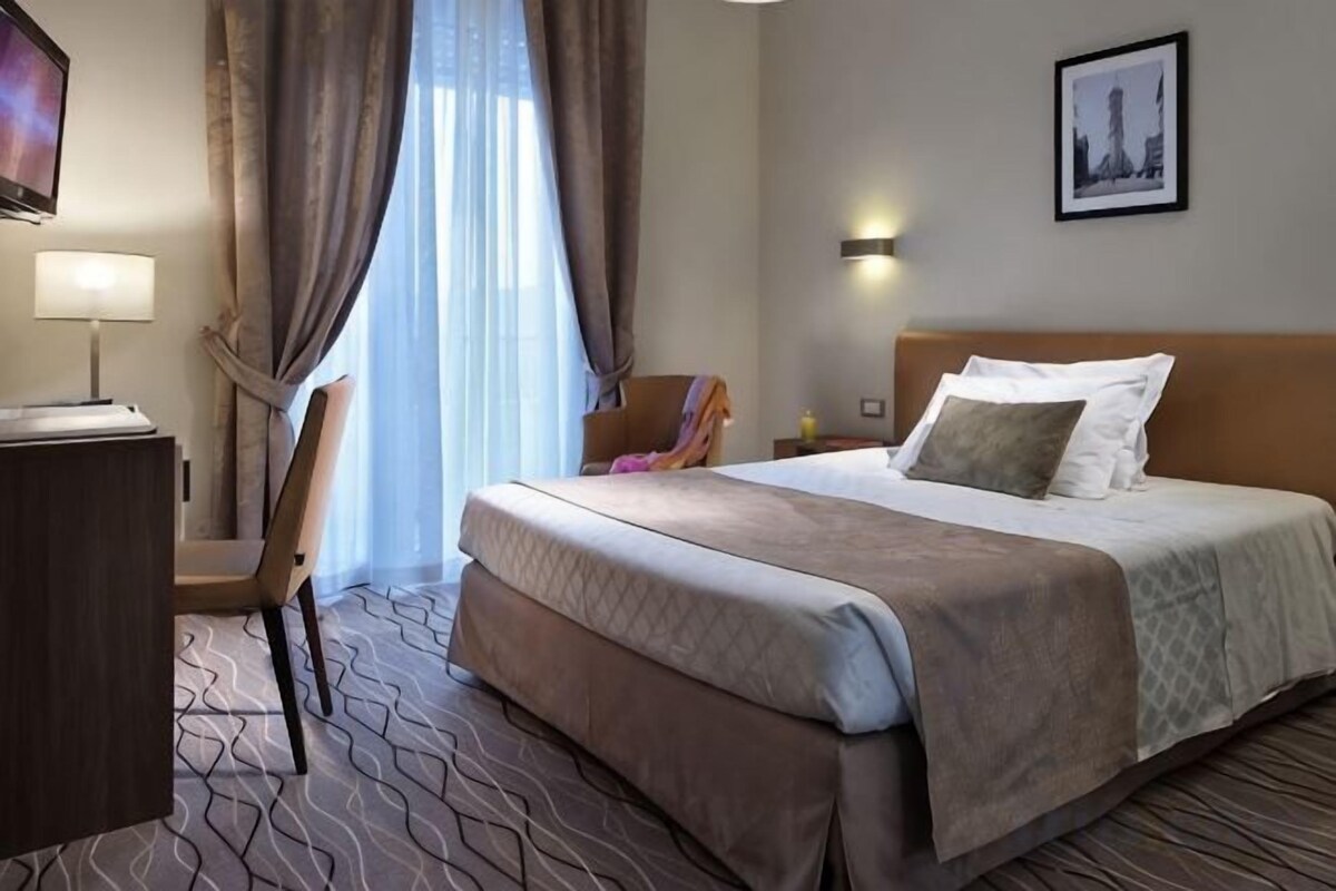 A hotel room with a bed, a desk, a chair, a lamp, and a picture on the wall.