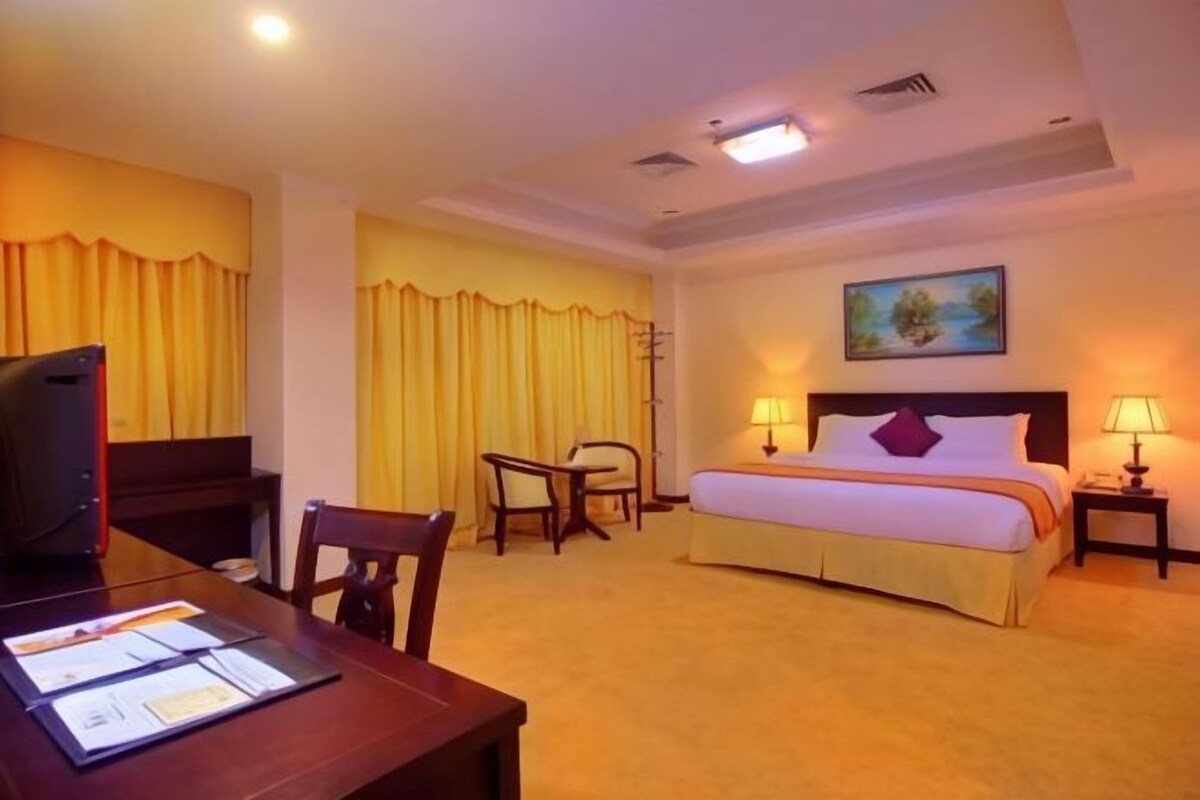 Ein Hotelzimmer mit Bett, Schreibtisch mit Unterlagen, Stuhl, Fernseher und einem Bild an der Wand.