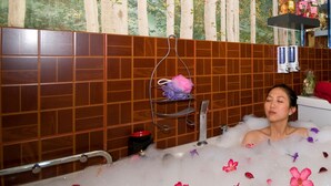 A bathtub filled with bubbles and rose petals, a metal rack with towels, and a tiled wall with a nature-themed design.
