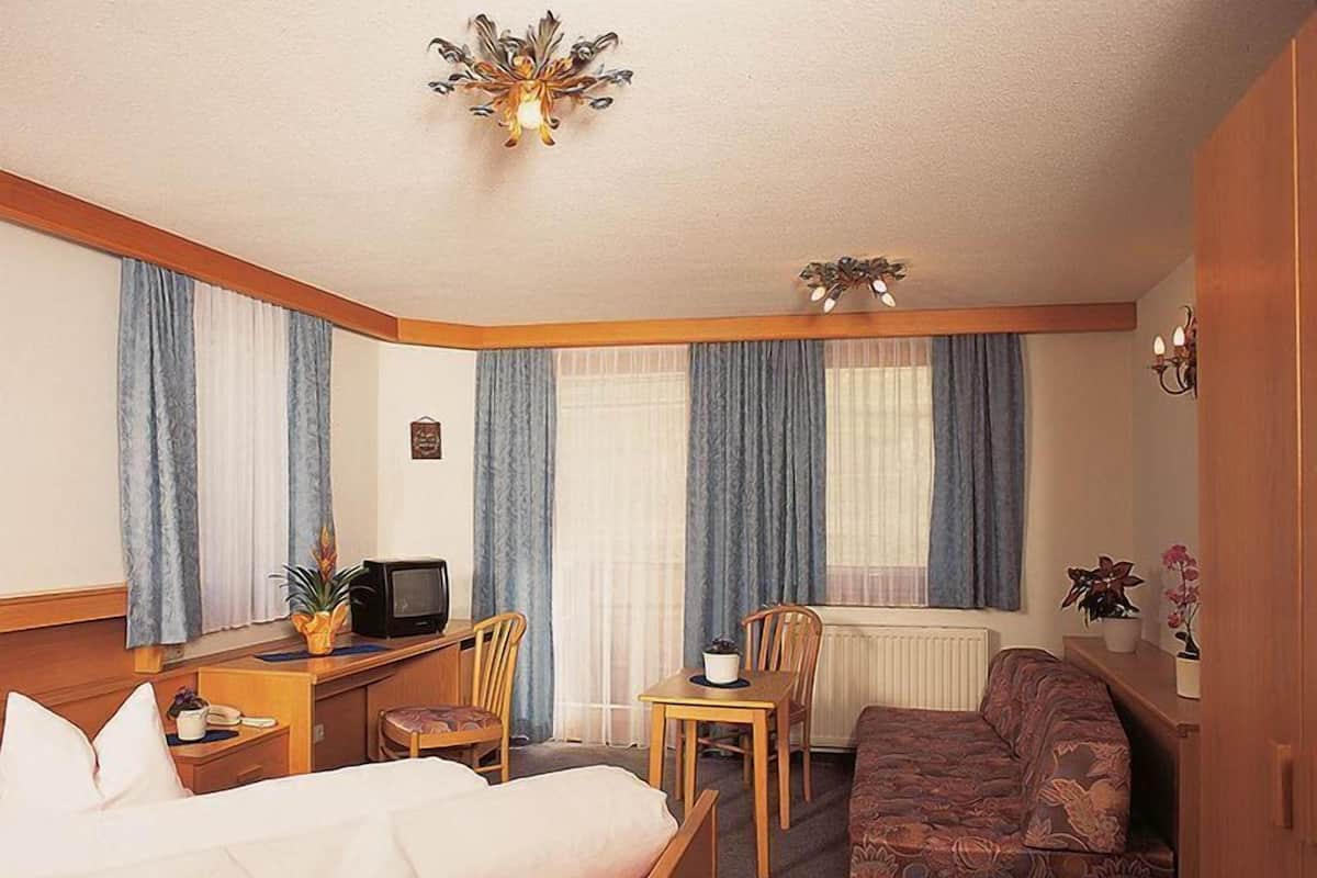 A hotel room with a bed, a desk with a computer, a chair, a sofa, and a window with curtains.