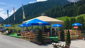 A mountain lodge with outdoor seating under blue umbrellas, surrounded by lush greenery and a mountain backdrop. - Gasthof Martha (Nauders)