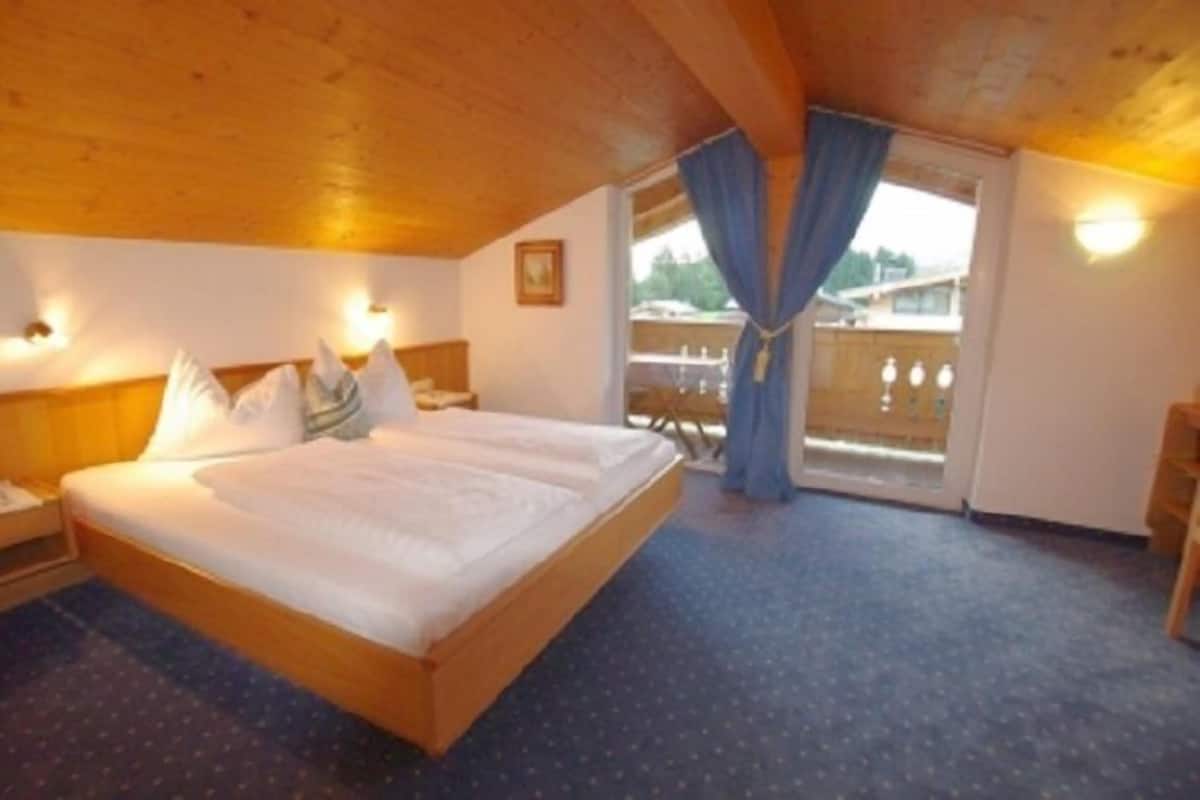 A hotel room with a wooden ceiling, a large bed with white linens, a nightstand, and a view of the outdoors through a balcony with blue curtains.