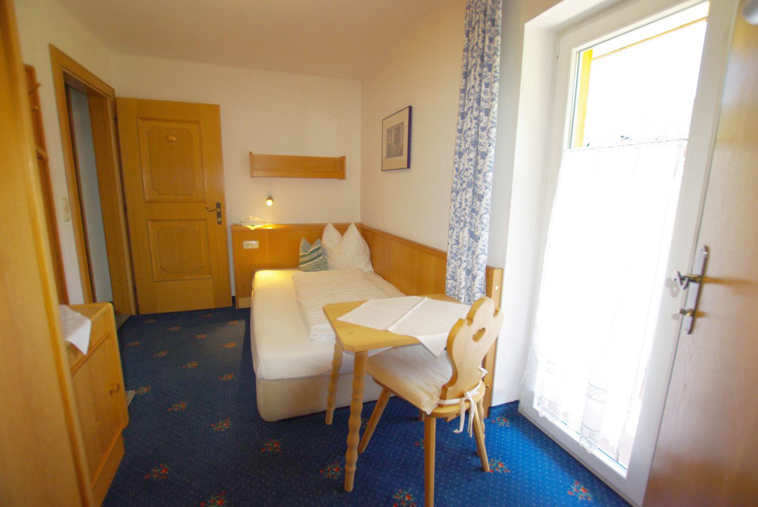 Habitación de hotel compacta con una cama individual, una mesita pequeña y una silla. Se ve una puerta a la izquierda, una ventana a la derecha y un estante fijado a la pared sobre la cama.