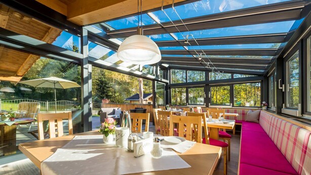 A spacious indoor area with wooden tables and chairs, large windows, and a glass ceiling allowing natural light. - Hotel Pension Heike (Aurach bei Kitzbuehel)