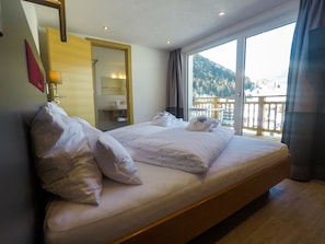 A hotel room with a large bed, a bathroom with a mirror, and a balcony with a view of the mountains. - Collina Hotel Garni (Nauders)