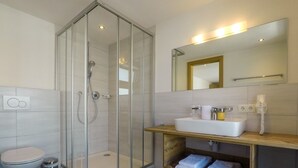A modern bathroom with a glass-enclosed shower, a white toilet, a sink with a mirror above it, and a shelf with folded towels. - Collina Hotel Garni (Nauders)