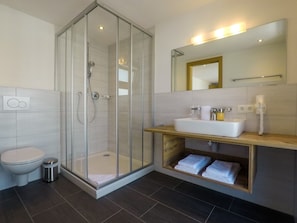 A modern bathroom with a glass-enclosed shower, a white toilet, a sink with a mirror above it, and a shelf with folded towels. - Collina Hotel Garni (Nauders)