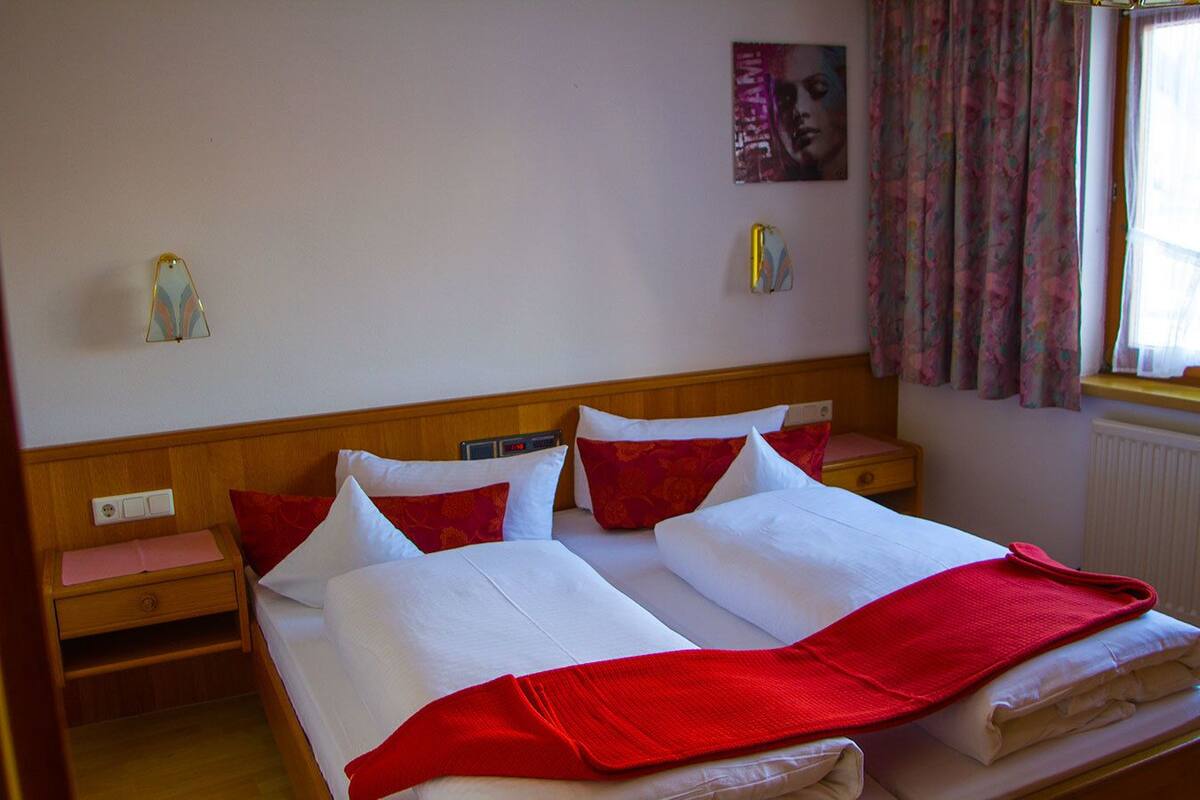 Ein Doppelbett mit weißer und roter Bettwäsche, zwei Nachttische, ein Wanduhren und ein Fenster mit Vorhängen.