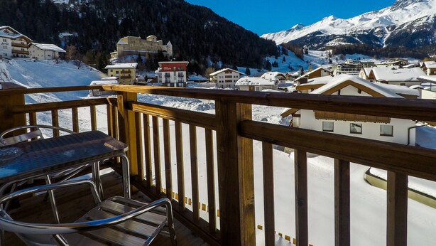 A wooden balcony with a metal chair, overlooking a snow-covered village with buildings and mountains in the background. - Collina Hotel Garni (Nauders)