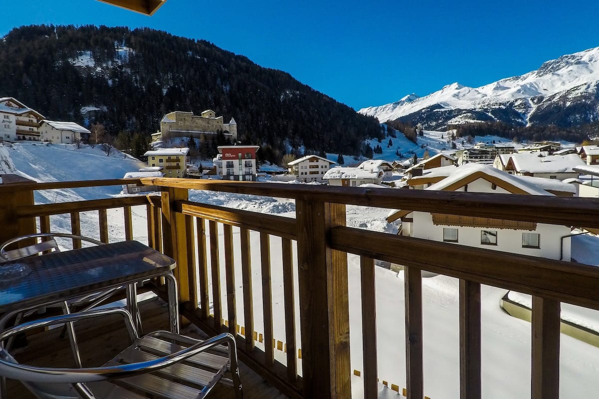 Ein hölzerner Balkon mit einem Metallstuhl, mit Blick auf ein verschneites Dorf mit Gebäuden und Bergen im Hintergrund.