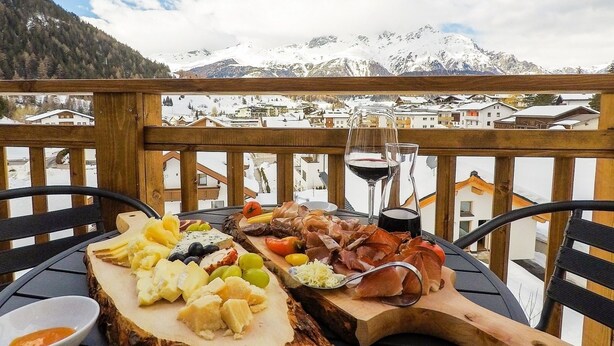 A wooden deck with a table set for a cheese and charcuterie board, a glass of red wine, and a scenic mountain view. - Collina Hotel Garni (Nauders)