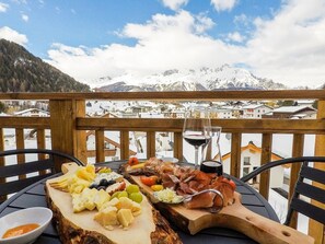 A wooden deck with a table set for a cheese and charcuterie board, a glass of red wine, and a scenic mountain view. - Collina Hotel Garni (Nauders)