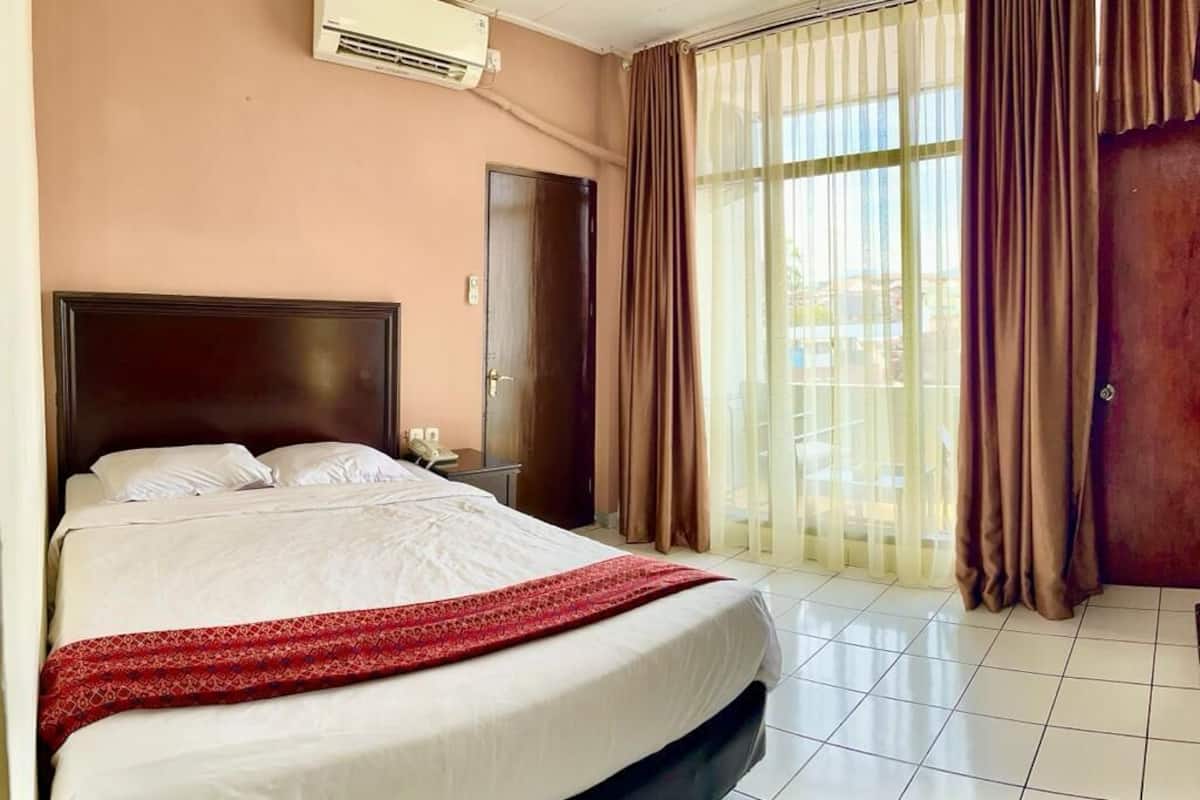 A hotel room with a bed, a nightstand, an air conditioning unit, a door, and a window with curtains.