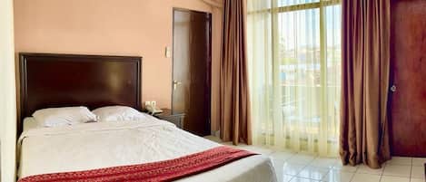 Une chambre d’hôtel comprenant un lit, une table de chevet, un appareil de climatisation, une porte et une fenêtre avec des rideaux.