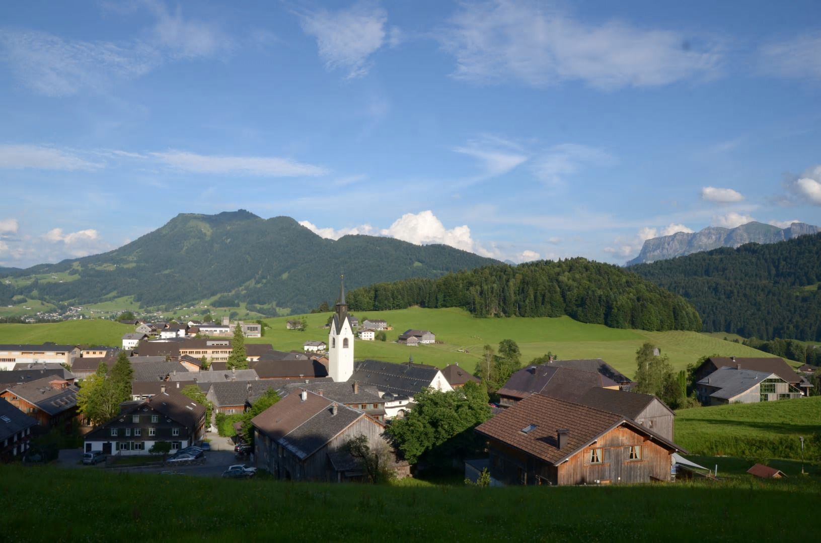 Ein ländliches Dorf mit einer markanten Kirche, umgeben von grünen Feldern und Bergen im Hintergrund.