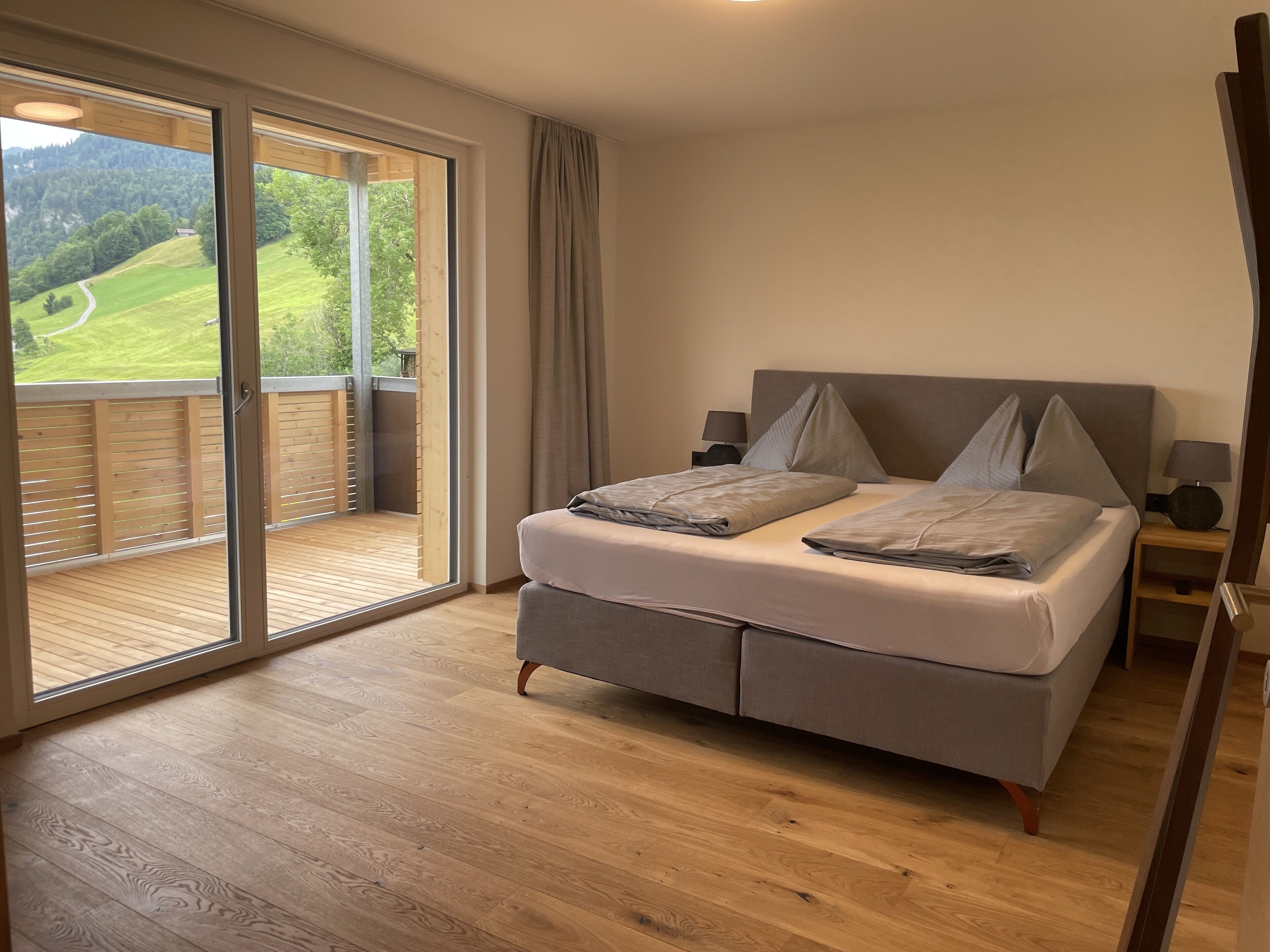 A bedroom with a large sliding glass door leading to a balcony, a bed with grey bedding, two bedside tables with lamps, and a view of a green landscape.
