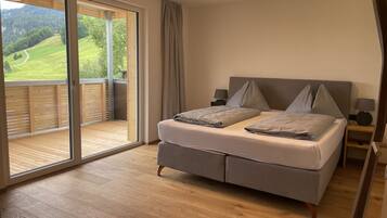 A bedroom with a large sliding glass door leading to a balcony, a bed with grey bedding, two bedside tables with lamps, and a view of a green landscape.