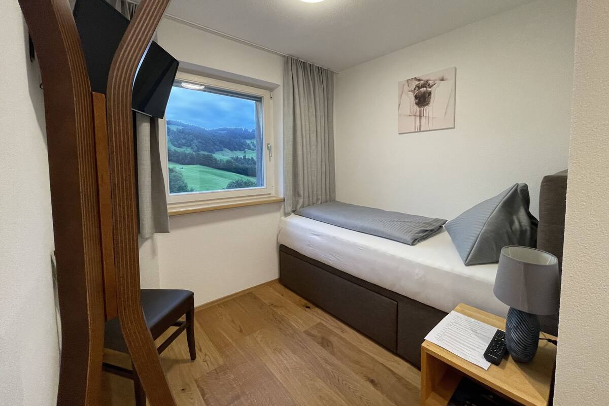 A small bedroom with a single bed, a window with a view of green fields, a desk with a lamp, and a chair.
