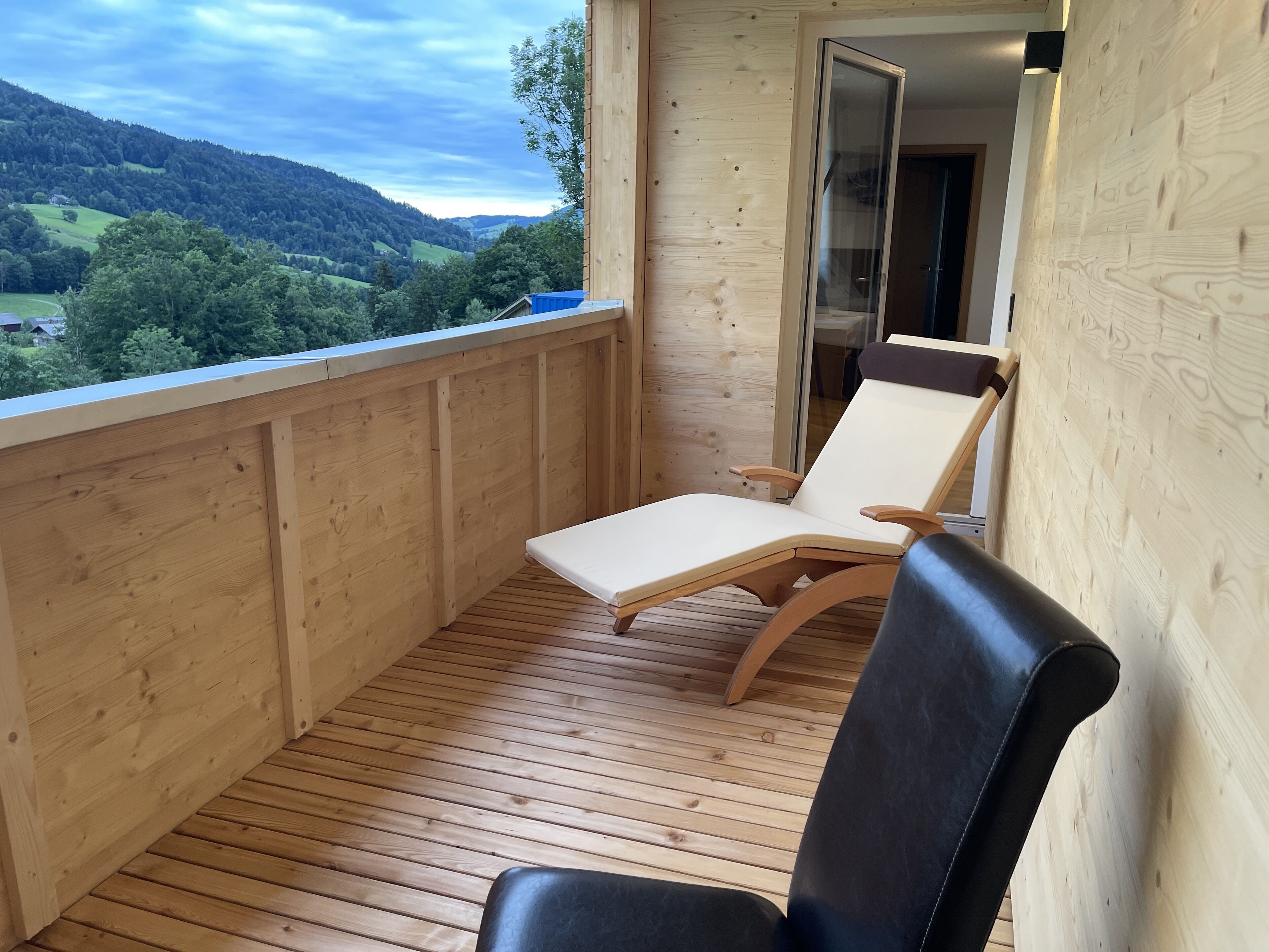 A wooden deck with two lounge chairs, one with a white cushion and the other with a black cushion, overlooking a scenic view of green hills and a clear sky.