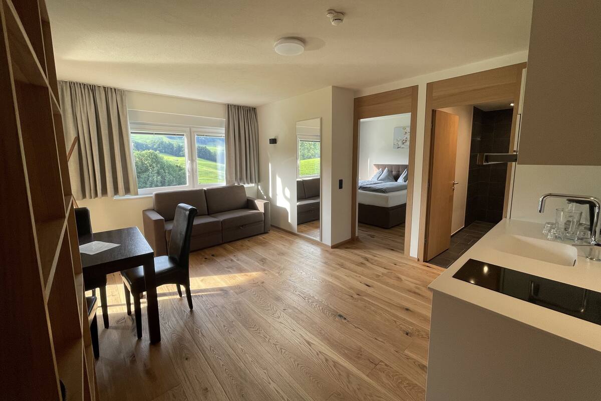 A modern, open-plan living space with a wooden floor, a sofa, a dining table with chairs, a kitchen area with a sink, and a view of a green landscape through large windows.