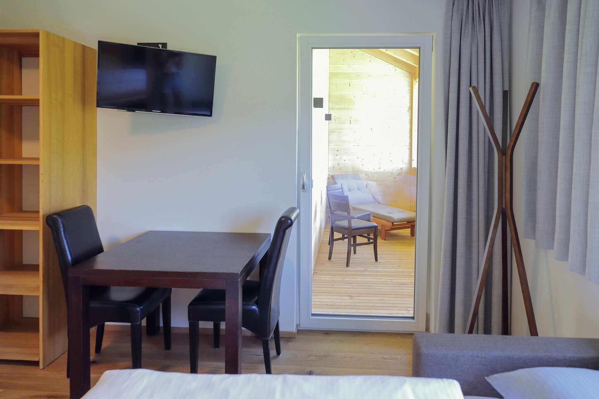 A room with a bed, a TV mounted on the wall, a wooden dining table with chairs, and a view of an outdoor balcony with a sofa.