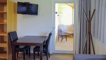 A room with a bed, a TV mounted on the wall, a wooden dining table with chairs, and a view of an outdoor balcony with a sofa.