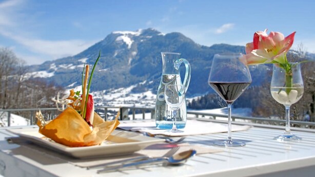 Una mesa dispuesta con un plato de comida, una jarrón de agua y dos copas de vino con flores, con vistas a un paisaje montañoso nevado.
