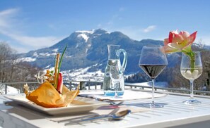 Una mesa dispuesta con un plato de comida, una jarrón de agua y dos copas de vino con flores, con vistas a un paisaje montañoso nevado.