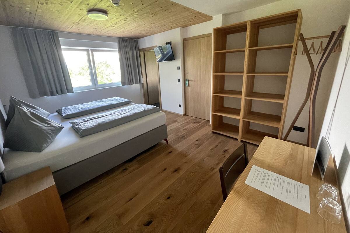 A bedroom with a bed, a wooden wardrobe, a desk with a chair, and a window with curtains.