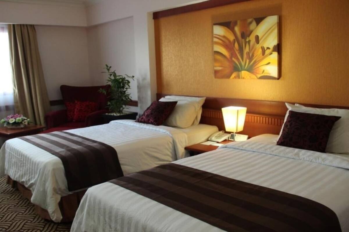 A hotel room with two beds, a nightstand with a lamp, a chair, a plant, and a painting on the wall.