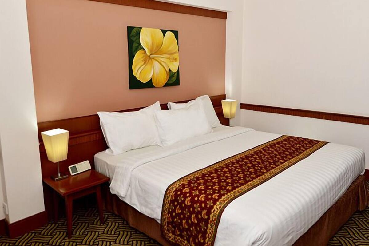 A hotel room with a large bed, two bedside tables, a nightstand, a lamp, a digital clock, and a floral painting on the wall.