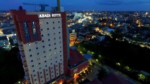 Un hôtel de grande hauteur appelé ABADI SUITE, avec de nombreux étages, une façade rouge et blanche bien visible et une vue sur le paysage urbain environnant au crépuscule.