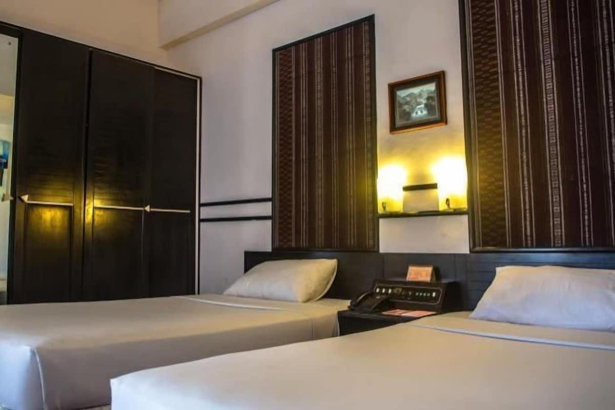 A hotel room with two beds, a wardrobe, and a bedside table with a lamp.