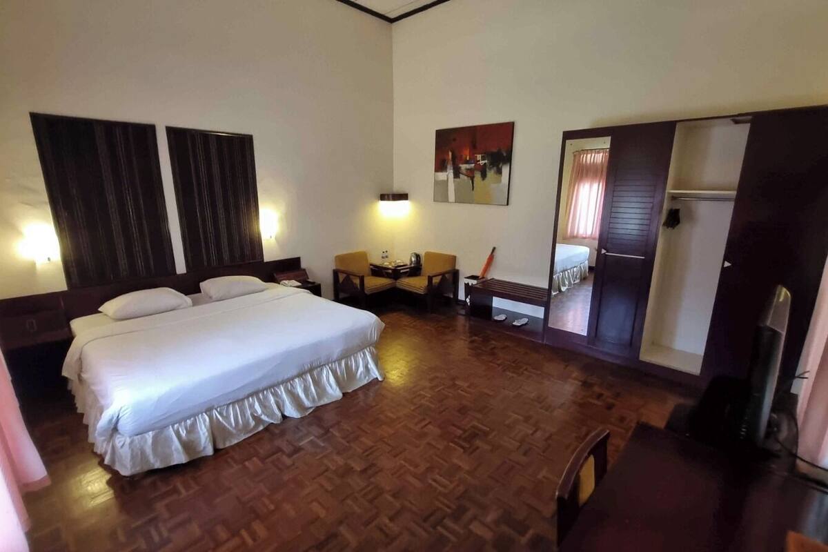 A hotel room with a large bed, a desk, a chair, a small table, a mirror, and a wardrobe.