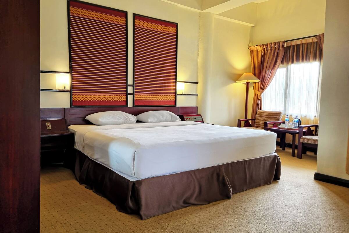 A hotel room with a large bed, two bedside tables, a nightstand, a desk with a chair, and a window with curtains.