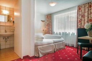 A hotel room with a bed, a small table, a chair, a window with curtains, a mirror, and a sink.