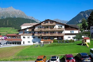 A multi-story hotel with balconies, a parking area, and a mountainous backdrop.