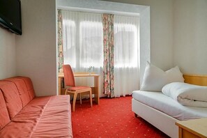 A hotel room with a red carpet, a white sofa, a bed with white pillows, a small table with a chair, and a window with floral curtains.