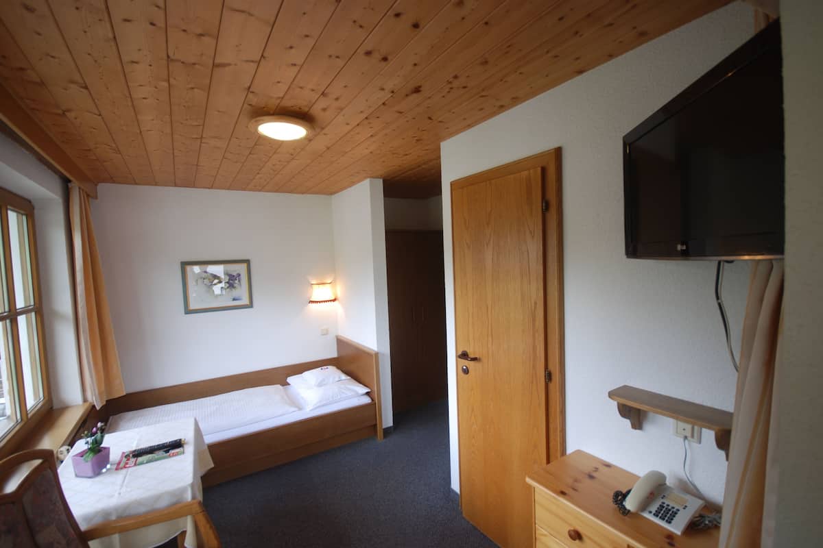 A small, cozy room with a wooden ceiling, a single bed, a desk with a chair, a television mounted on the wall, and a door leading to another room.