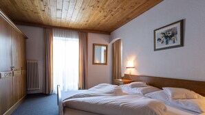 A hotel room with a wooden ceiling, a bed with white linens, a nightstand with a lamp, a radiator, and a window with curtains. - Pension Tirol (Nauders)