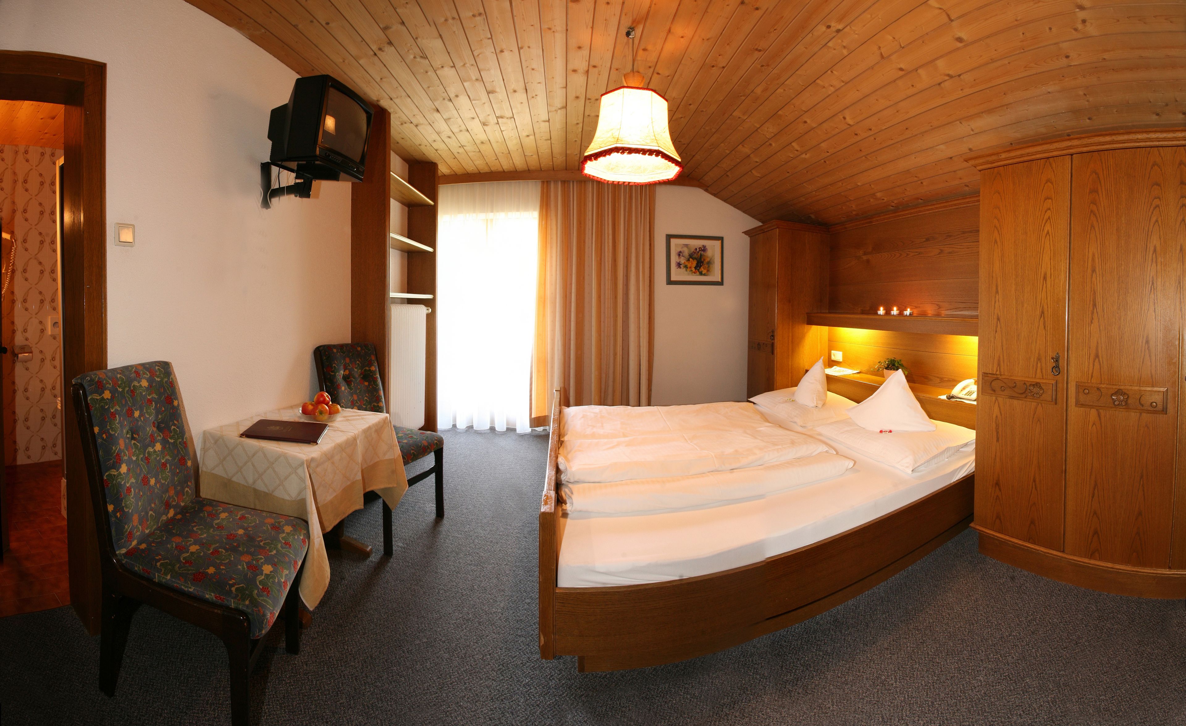 Un dormitorio con techo de madera, una cama con ropa de cama blanca, una silla con estampado floral y una mesita con un libro y fruta.