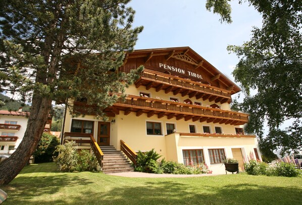 Un edificio de varios pisos con un cartel que dice "Pension Tirol". Cuenta con un balcón de madera en el piso superior, una escalera que conduce a la entrada y un césped bien cuidado al frente.