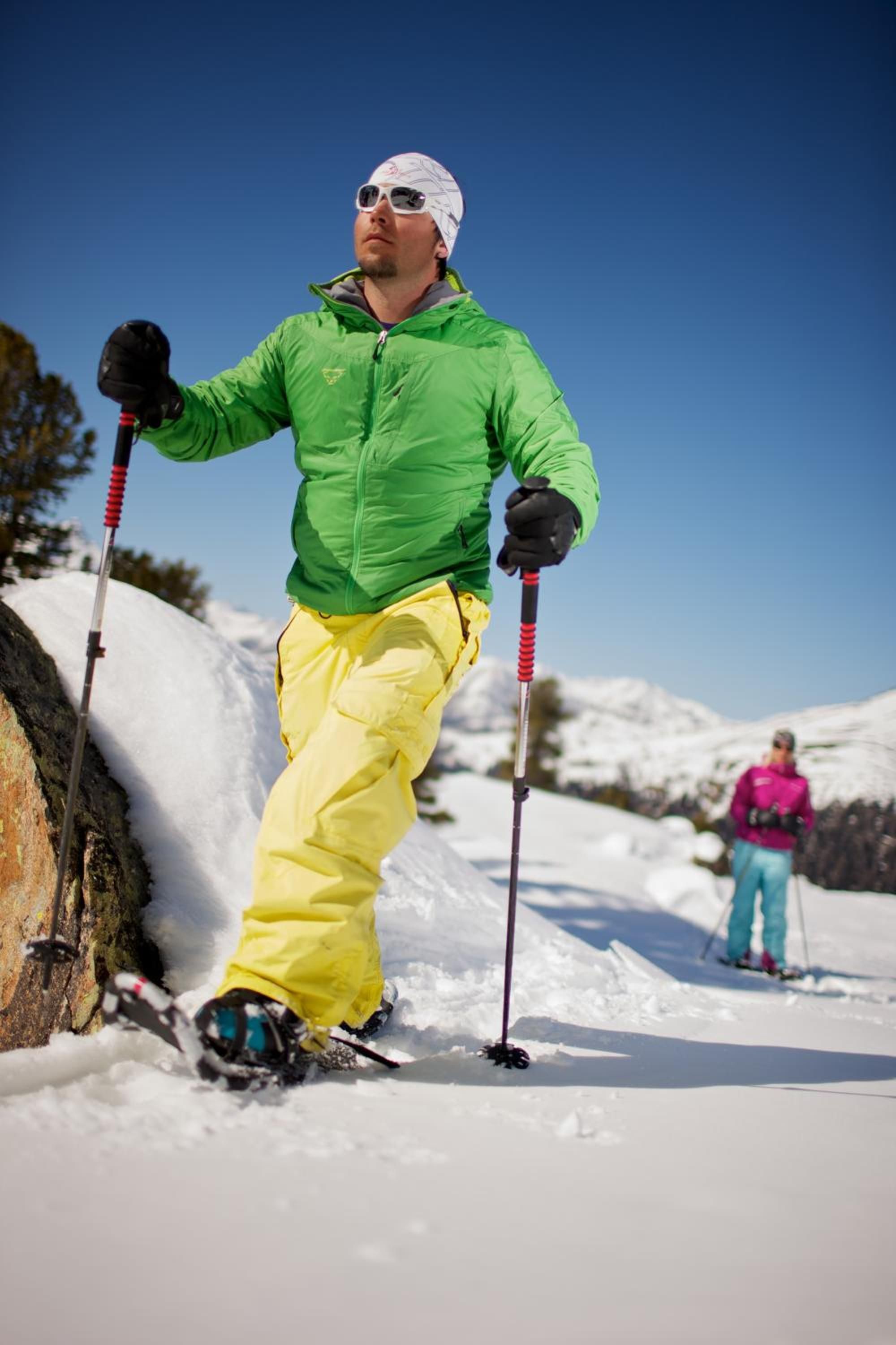 Eine Person mit Skistöcken fährt auf einem verschneiten Hang Ski, dabei trägt sie eine grüne Jacke und eine gelbe Hose.