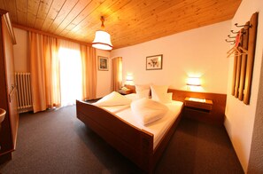Una cama con estructura de madera y sábanas blancas, una mesita de noche, un perchero de pared y un ventanal con cortinas.