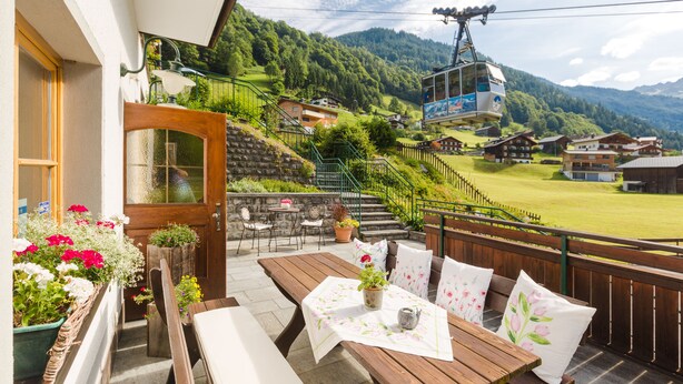 Una terraza de madera con una mesa, bancos, plantas en macetas y vistas a un pueblo de montaña con casas y un teleférico.