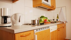 Una cocina con armarios de madera, encimera blanca, cafetera, hervidor, olla, verduras y una botella de vino.