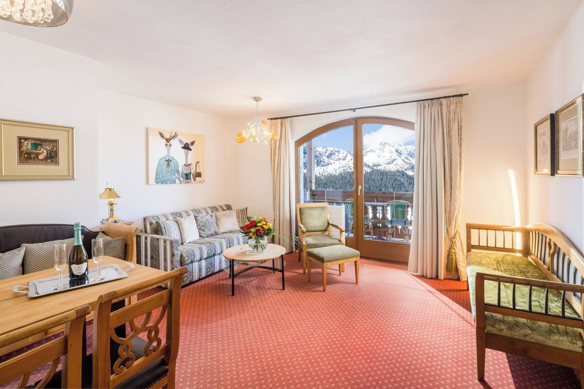 Una sala de estar acogedora con un sofá, sillas, una mesa de comedor con una botella y vasos, un florero con flores y un ventanal con vistas a la montaña.