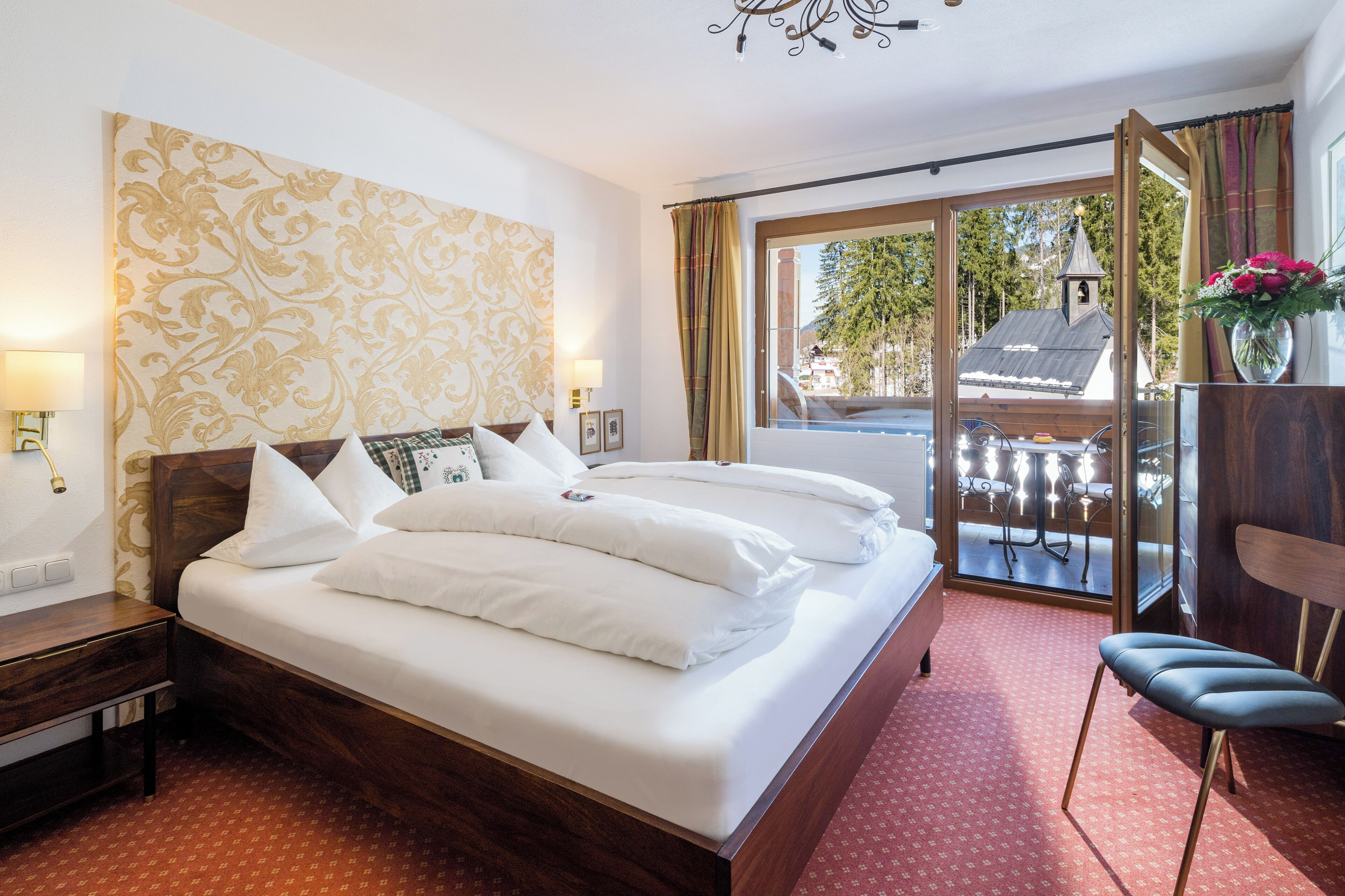 A hotel room with a large bed, a wooden headboard, a nightstand, a desk, a chair, a window with a view of a building and trees, and a balcony with a railing.